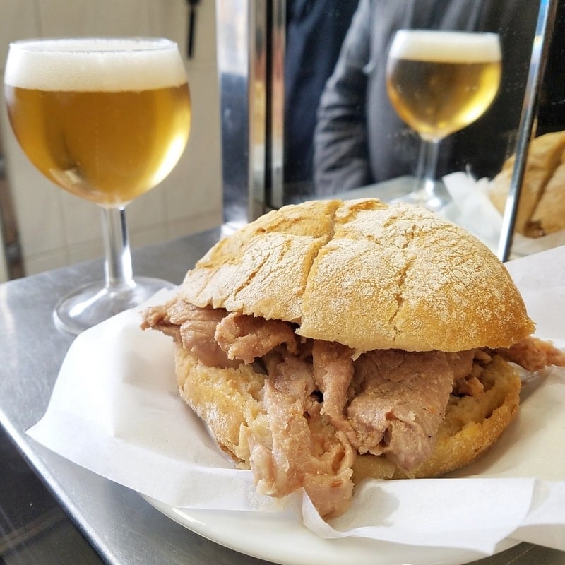 Close-up of a bifana pork sandwich on a table with two glasses of beer behind it.