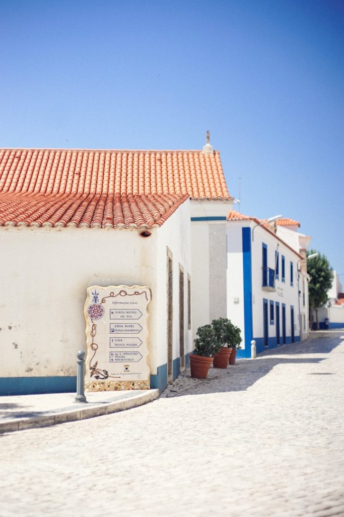 Sunny cobblestone street lined with blue-and-white buildings with tiled roofs and a painted sign. 