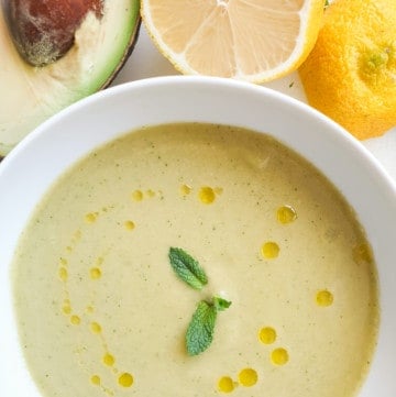 Green gazpacho in a white bowl with avocado and lemon in the background.