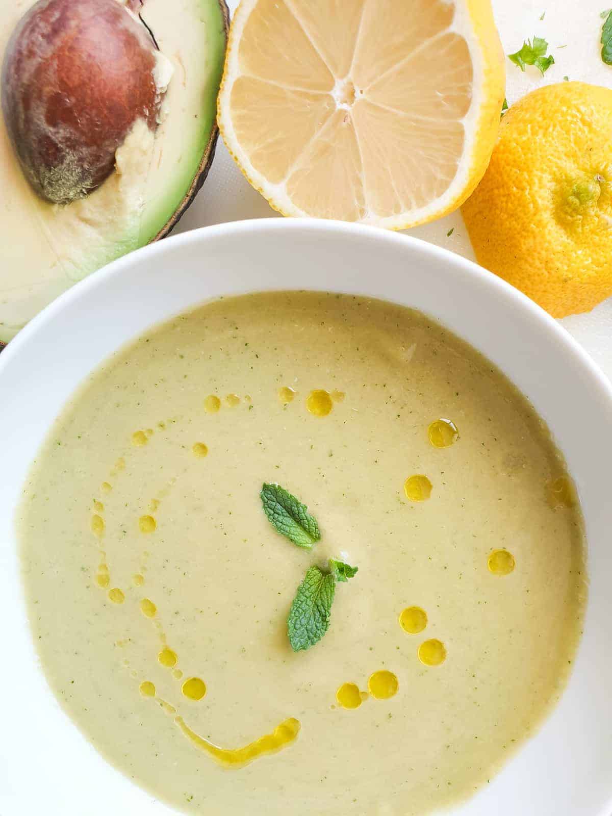 Green gazpacho in a white bowl with avocado and lemon in the background.