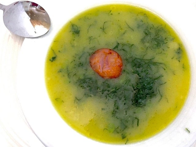 A bowl of caldo verde from above: yellow soup with shredded kale and a  sausage slice.