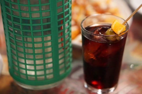 A tall glass of dark reddish brown vermouth with a skewered olive and orange slice.
