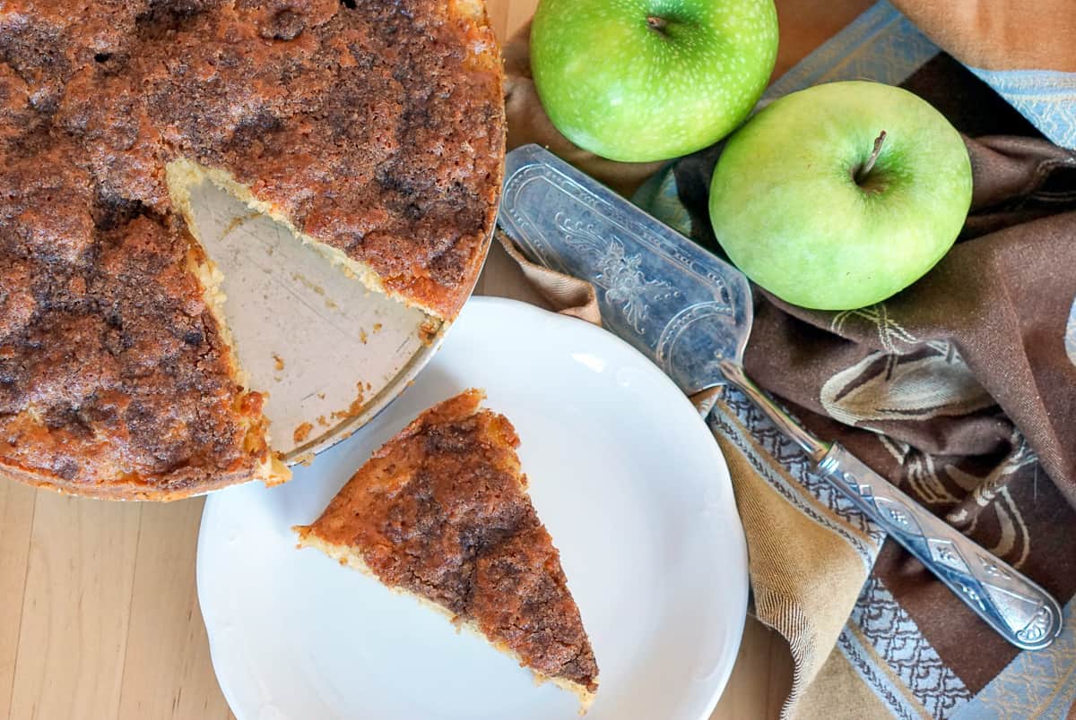 Apple Olive Oil Cake with one slice on a white plate and the whole cake in the background. Two granny smith apples on the side.