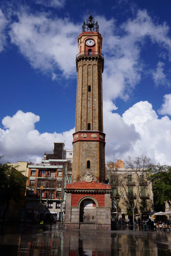 A tall clock tower made of gray and red stone in the center of a city square.