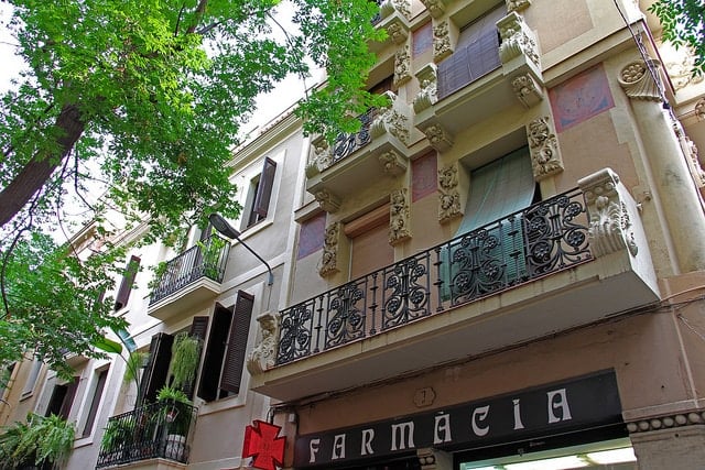 Pastel-colored building with an ornate iron balcony and molding, with a leafy tree beside it.