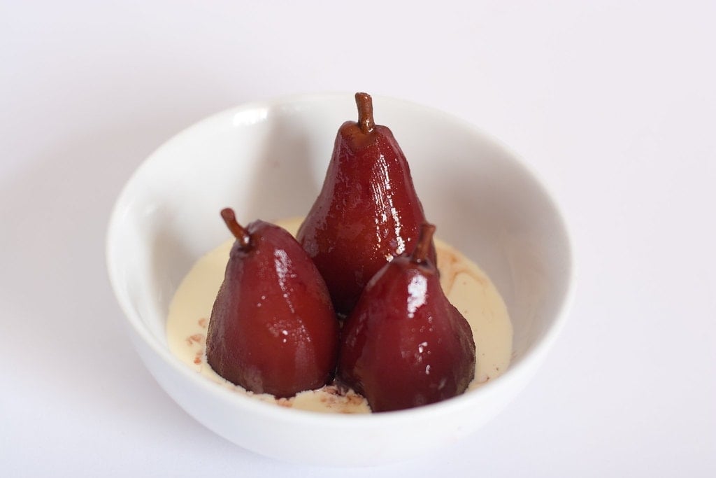 Three dark red pears poached in red wine and spices in a bowl of cream.