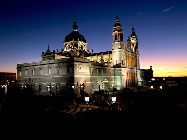 The Almudena Cathedral illuminated at night with a purple, pink, and yellow sunset behind it.