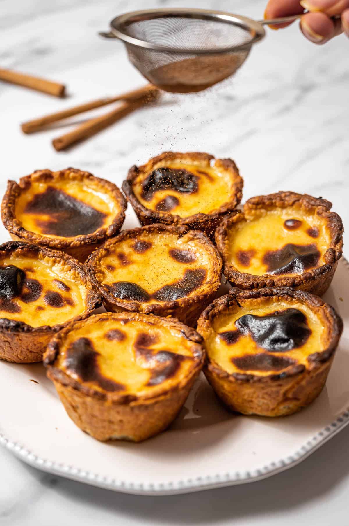 A plate of pasteis de nata (Portuguese custard tarts) with a sifter of ground cinnamon above