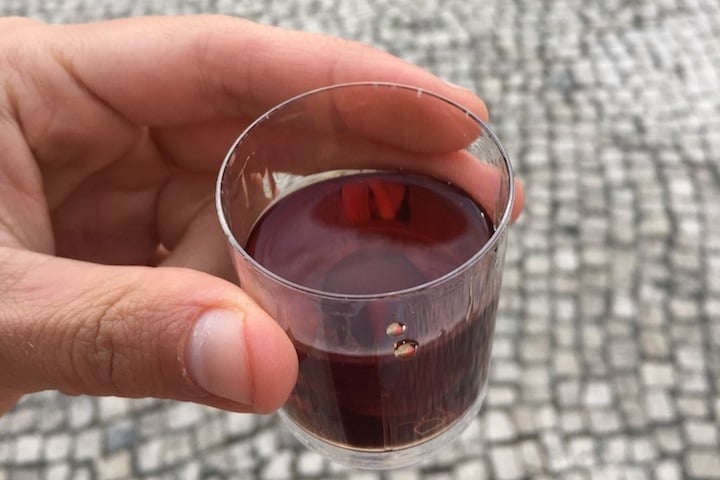 Close-up of a shot-sized glass of dark red liquid, with a tiled sidewalk in the background.