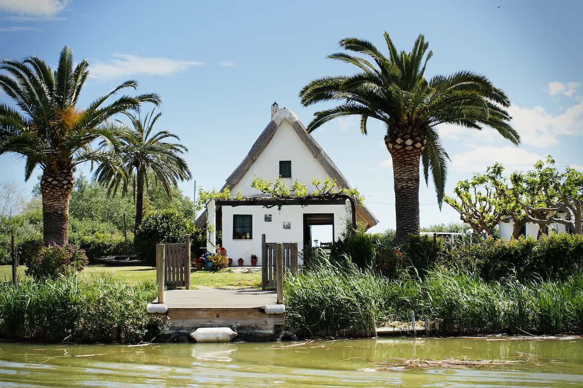 Small white house on a lake surrounded by palm trees and other plants.