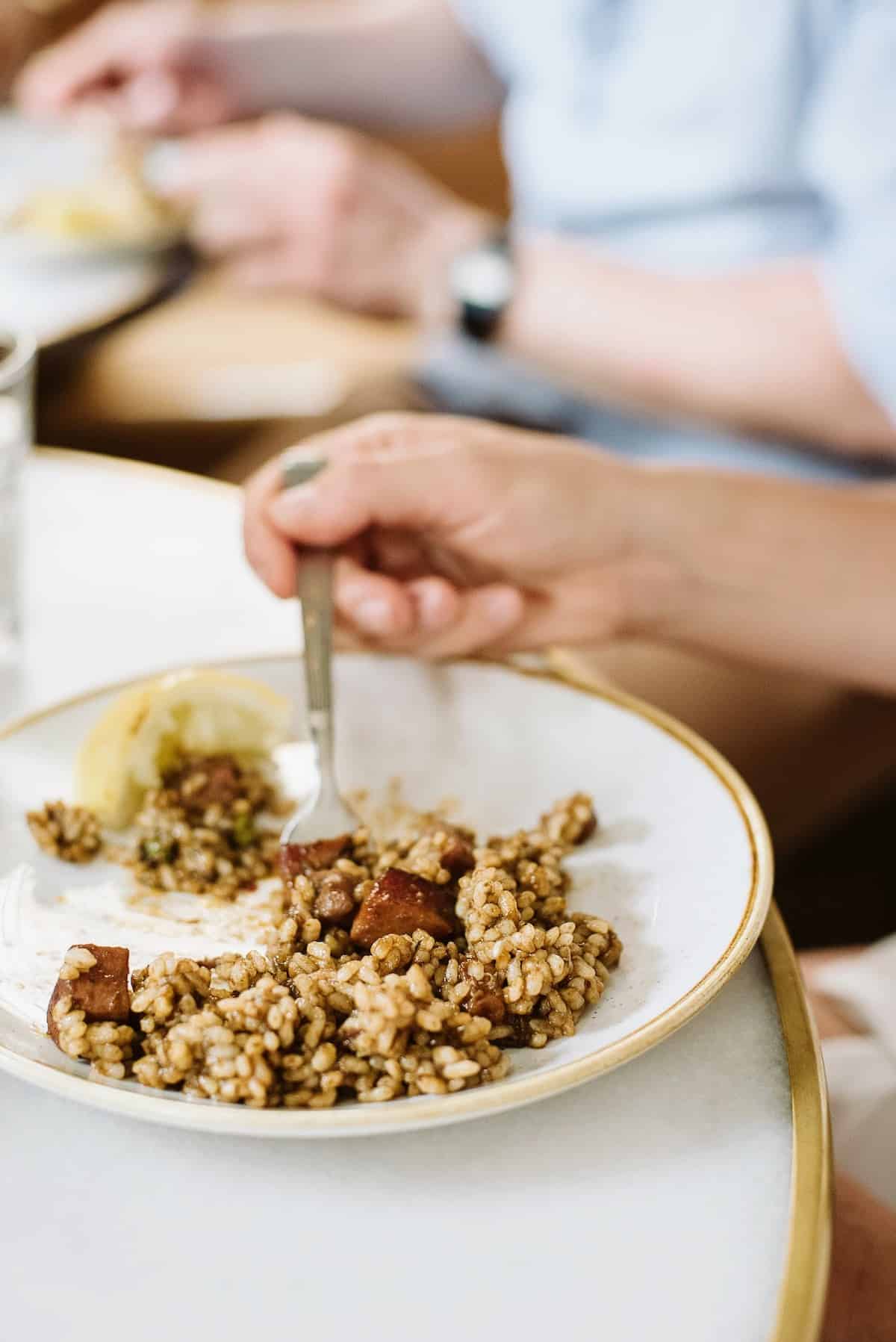 Person's hand eating a brown-colored seafood rice dish with a fork off a white plate.
