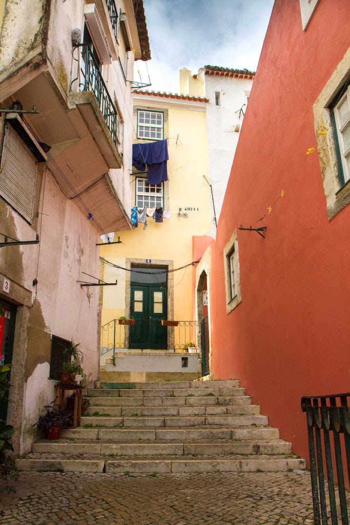 A narrow street in Alfama with stone stairs and orange, pink, and yellow buildings.