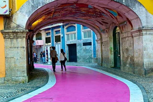 Two people walking down a pedestrian street painted bright pink, curving beneath an archway.