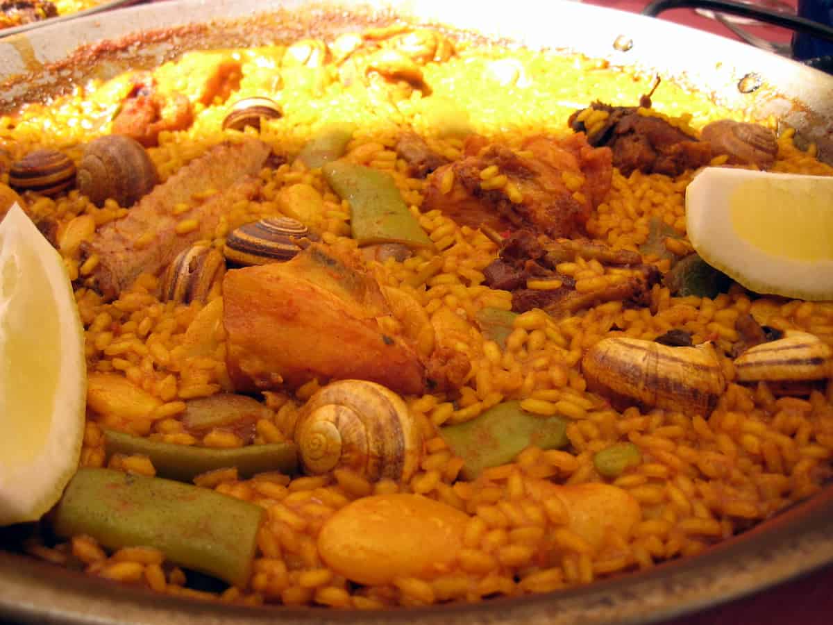 Close up of a pan of paella with lemon wedges, meat, beans, and snails.