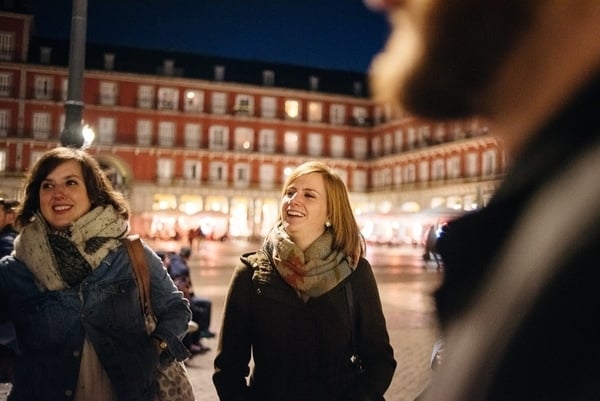 Two smiling women wearing scarves standing in Madrid's Plaza Mayor at night.