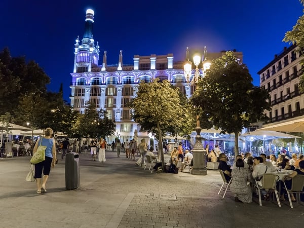 Plaza Santa Ana at night, with an ornate building illuminated in purple and several outdoor tables.