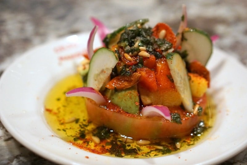 Close-up of a thick tomato slice topped with colorful fruits, vegetables, garnishes, and olive oil.