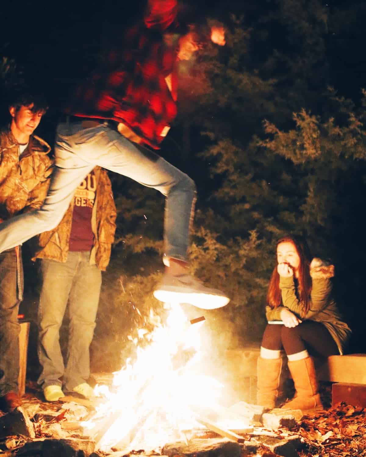 A man in a plaid shirt leaping over a bonfire while others look on