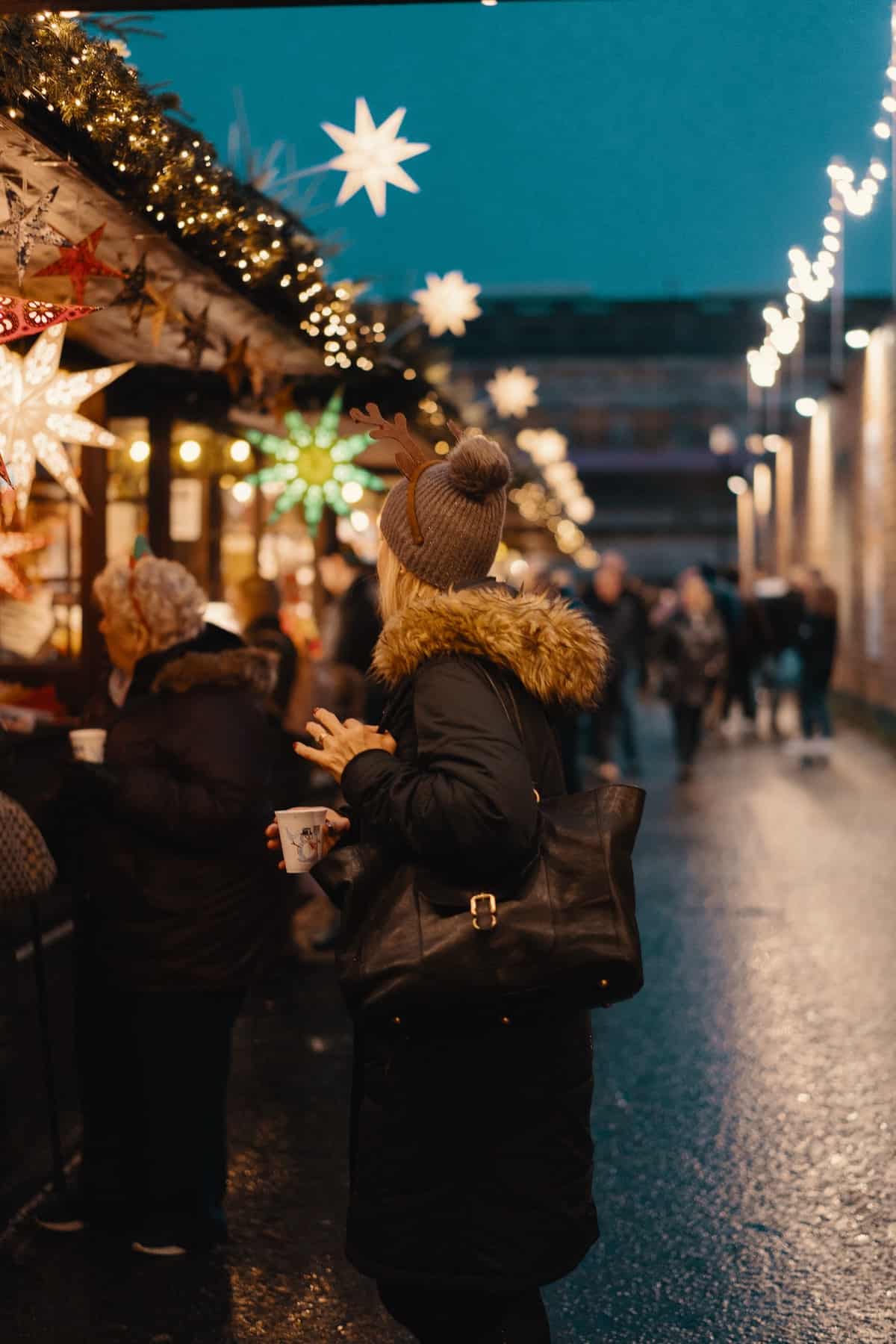 A woman standing among stalls at a brightly lit Christmas market.