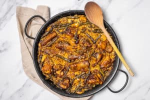 Overhead shot of a traditional Spanish paella with chicken and ribs in a black paella pan with a wooden spoon.
