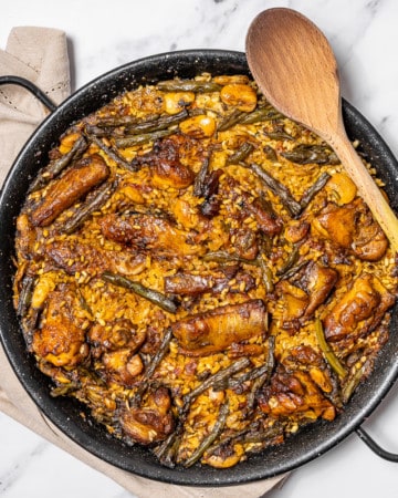 Overhead shot of a traditional Spanish paella with chicken and ribs in a black paella pan with a wooden spoon.