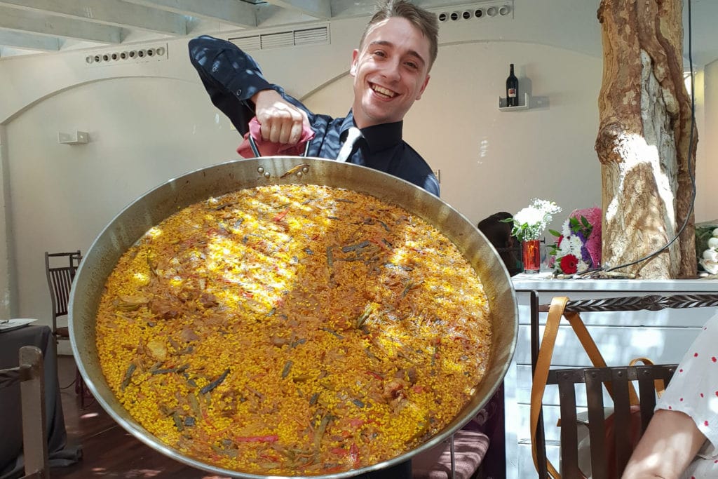 A grinning waiter holding up an enormous pan of bright yellow paella at a restaurant. 