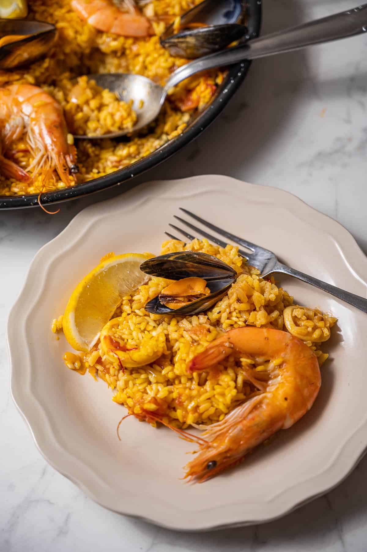 Serving of seafood paella on a white plate in front of the rest of the paella in the pan