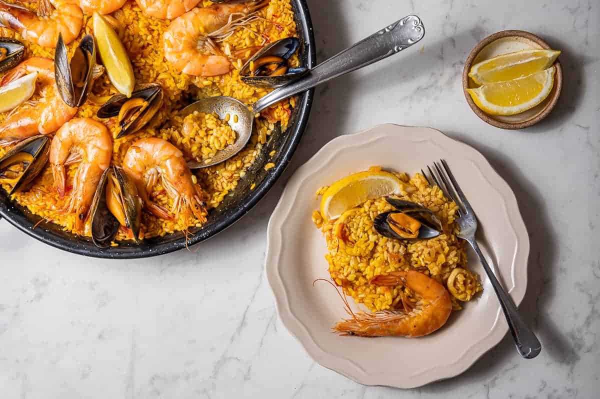 One serving of paella on a white plate with the rest of the paella pan in the background and some lemon wedges on the side.