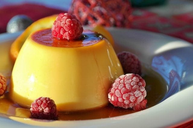 Yellow flan on a plate, sitting in a pool of caramel and garnished with raspberries.