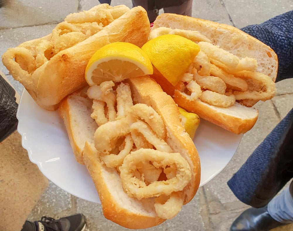 A white plate with three bread rolls stuffed with fried calamari and two lemon wedges.