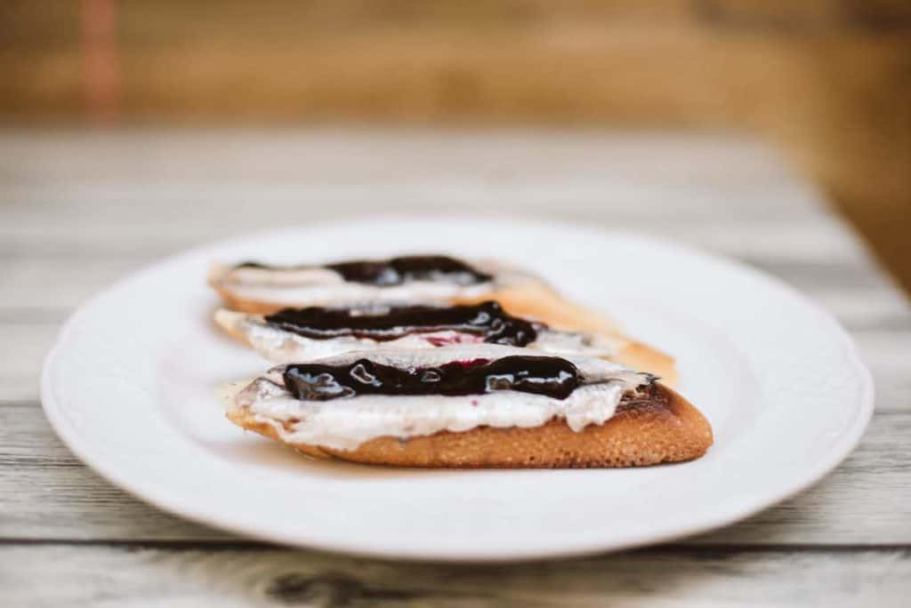 Three small pieces of bread topped with anchovies and blueberry jam on a white plate.