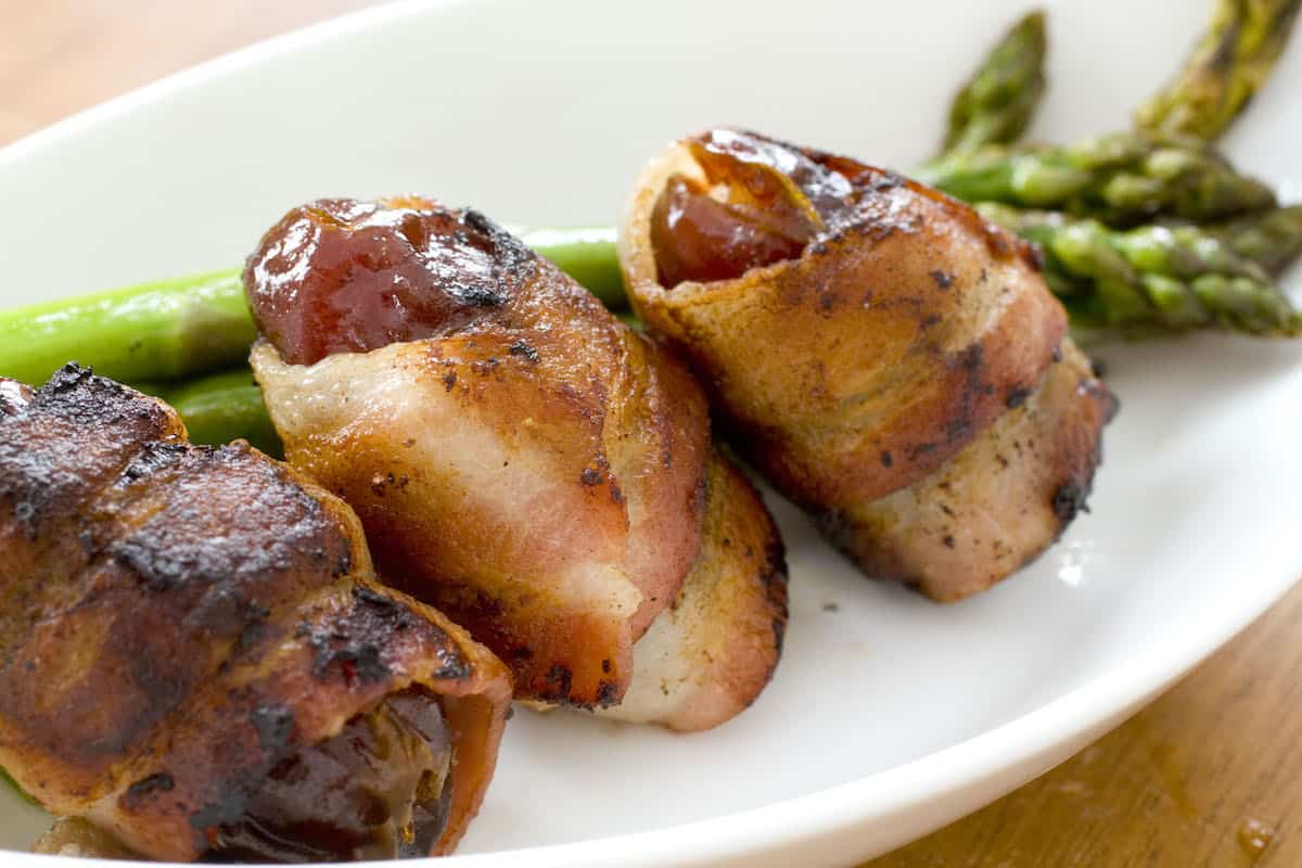 Three bacon-wrapped dates on a white plate in front of green asparagus.