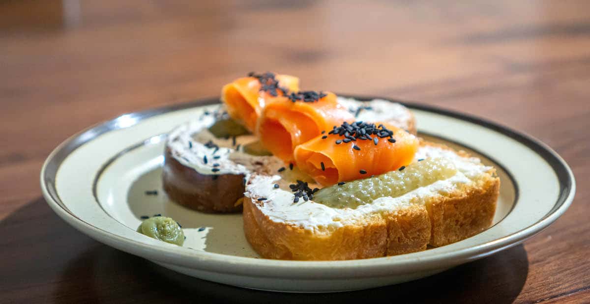 Slice of bread topped with cream cheese, smoked salmon, black sesame seeds, and pickles.