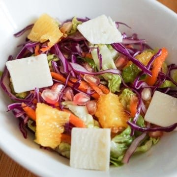 Winer salad with lettuce, cabbage carrot, pomegranate, orange and cheese in a white bowl.