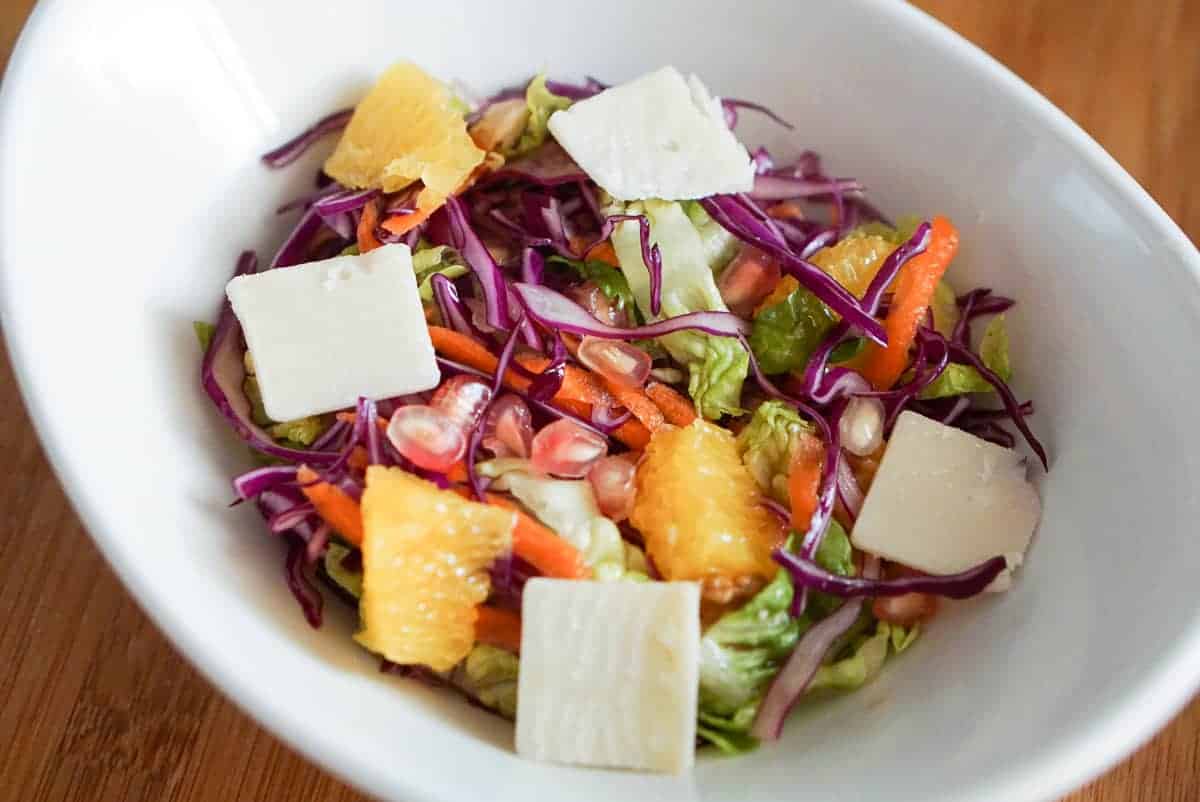 Winter salad with lettuce, cabbage, carrot, pomegranate, orange, and cheese in a white bowl.