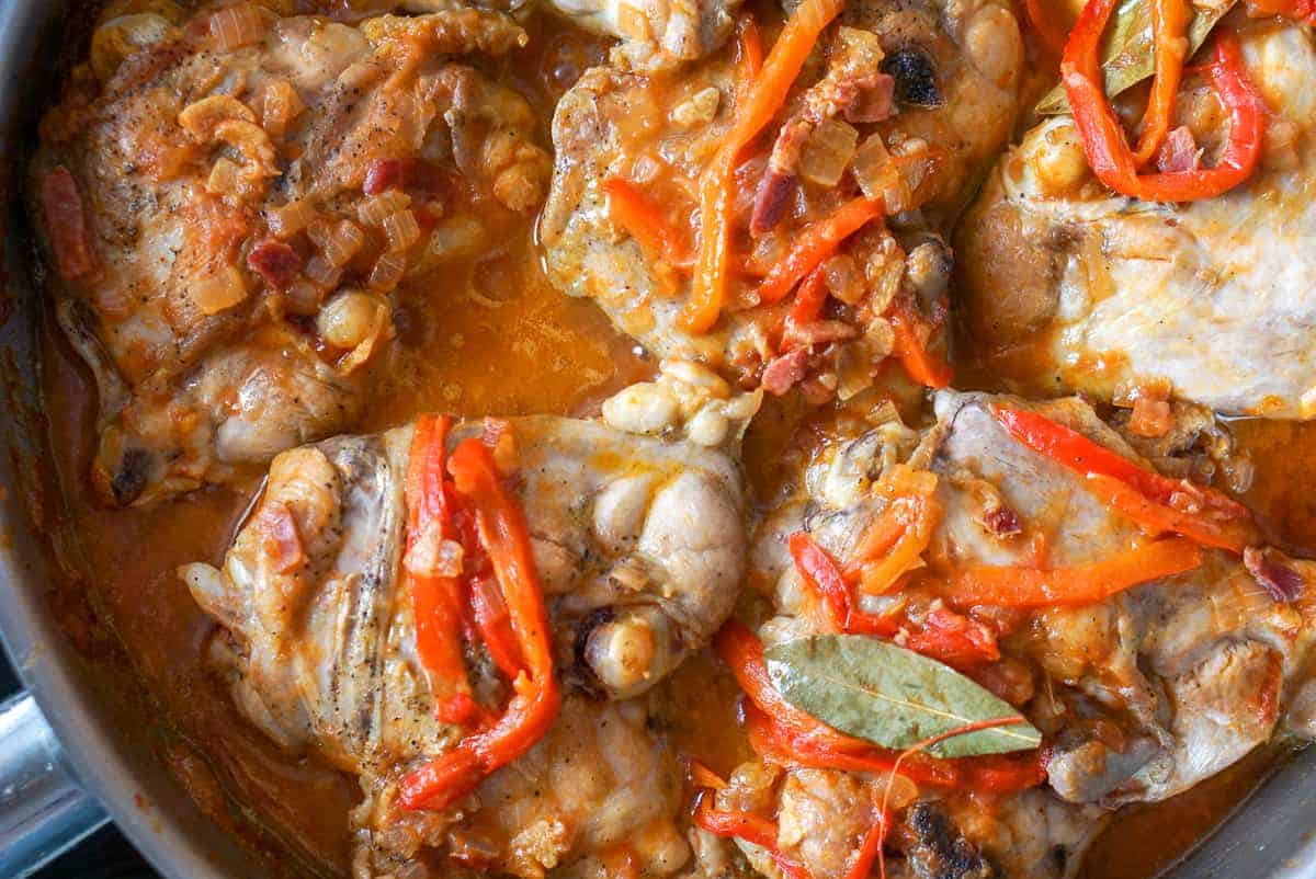 Chicken chilindron in a stainless steel skillet. An overhead photo of chicken thighs in a red sauce with onion and strips of bell pepper.