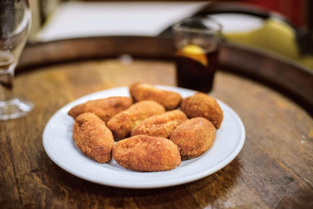 Spanish ham croquettes on a white plate with a glass of vermouth in the background.