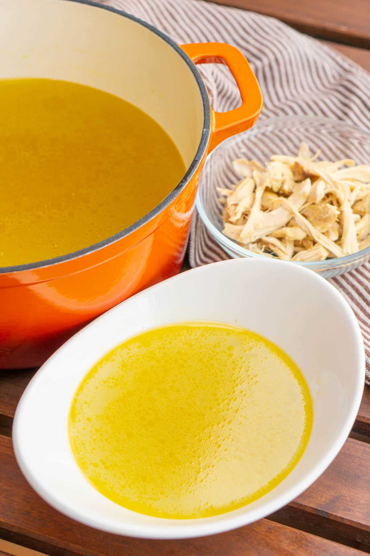 Spanish chicken broth in an orange Dutch oven pot in the background and a white bowl of broth at the front. A bowl of shredded chicken behind.