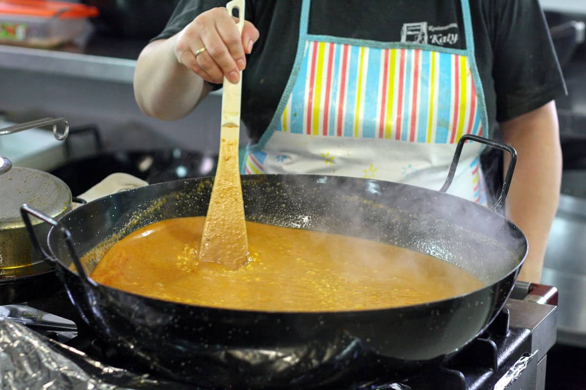 A woman wearing an apron stirring soupy rice in a large round pan.