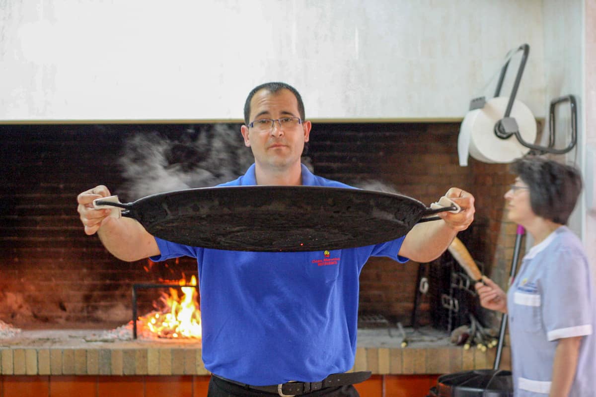 Man holding a large black paella pan in front of a fire in a brick oven.