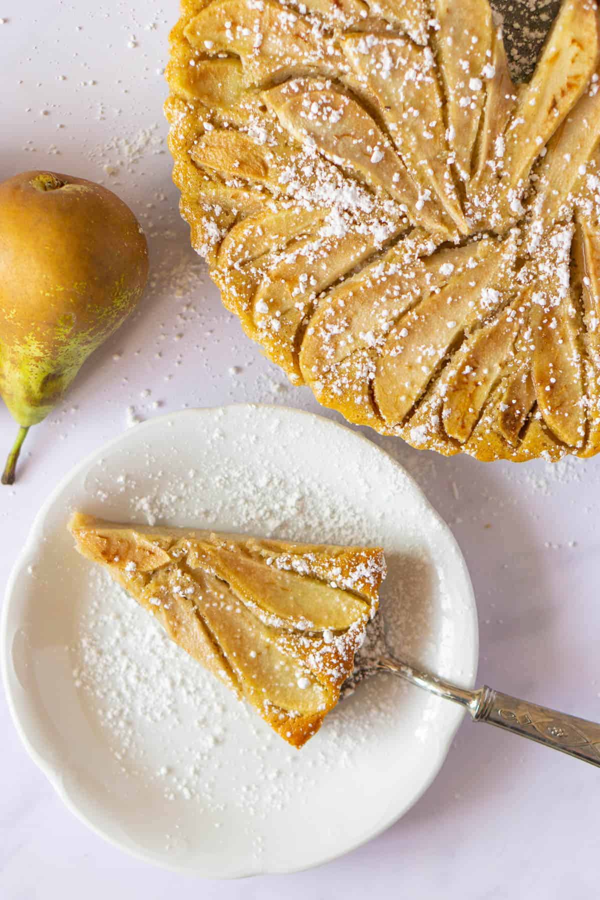 Pear tart slice on a white dish with rest of tart in the background and a pear on the side.