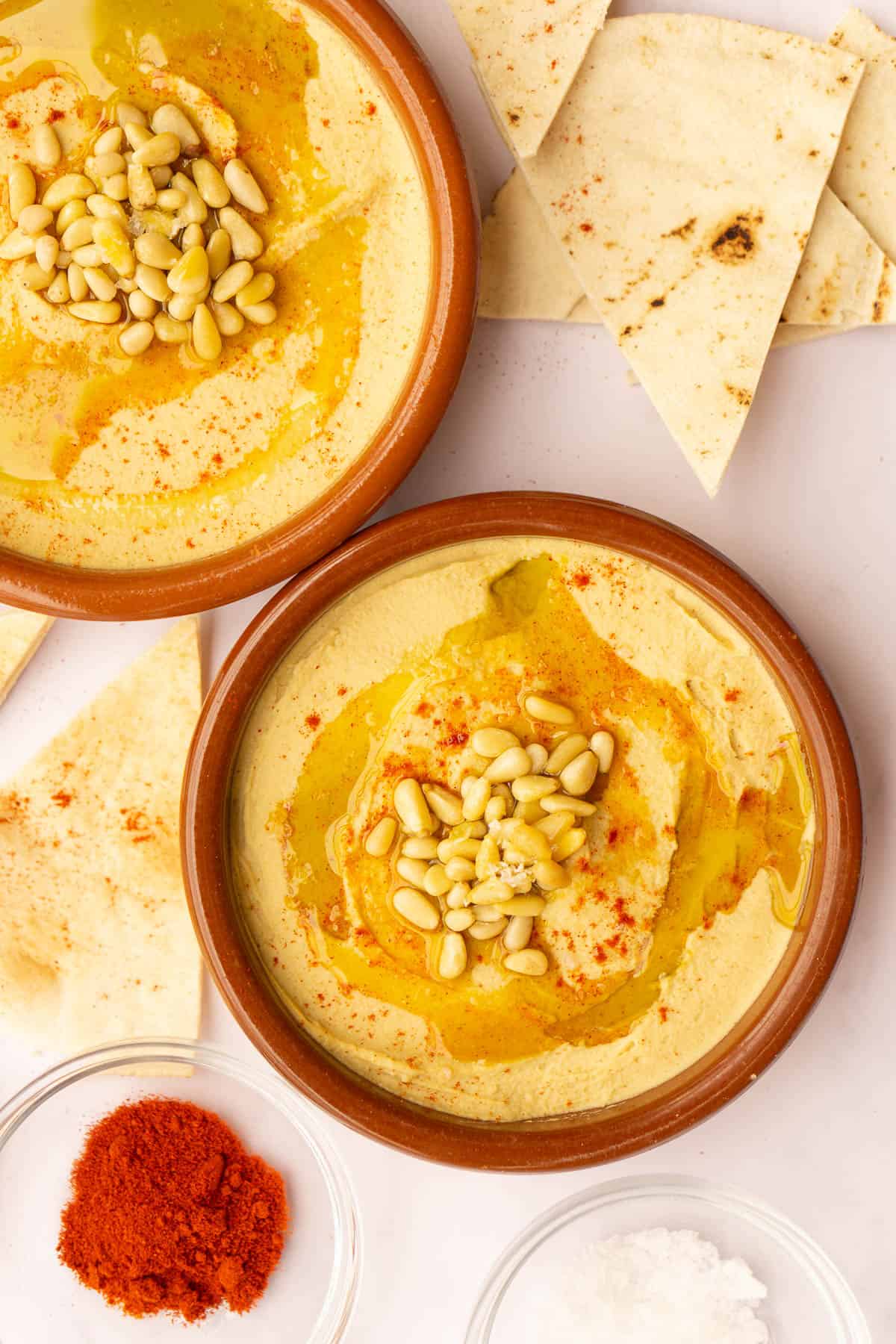 Two clay bowls of hummus topped with pine nuts, olive oil, and paprika. Pita bread and paprika in the background.