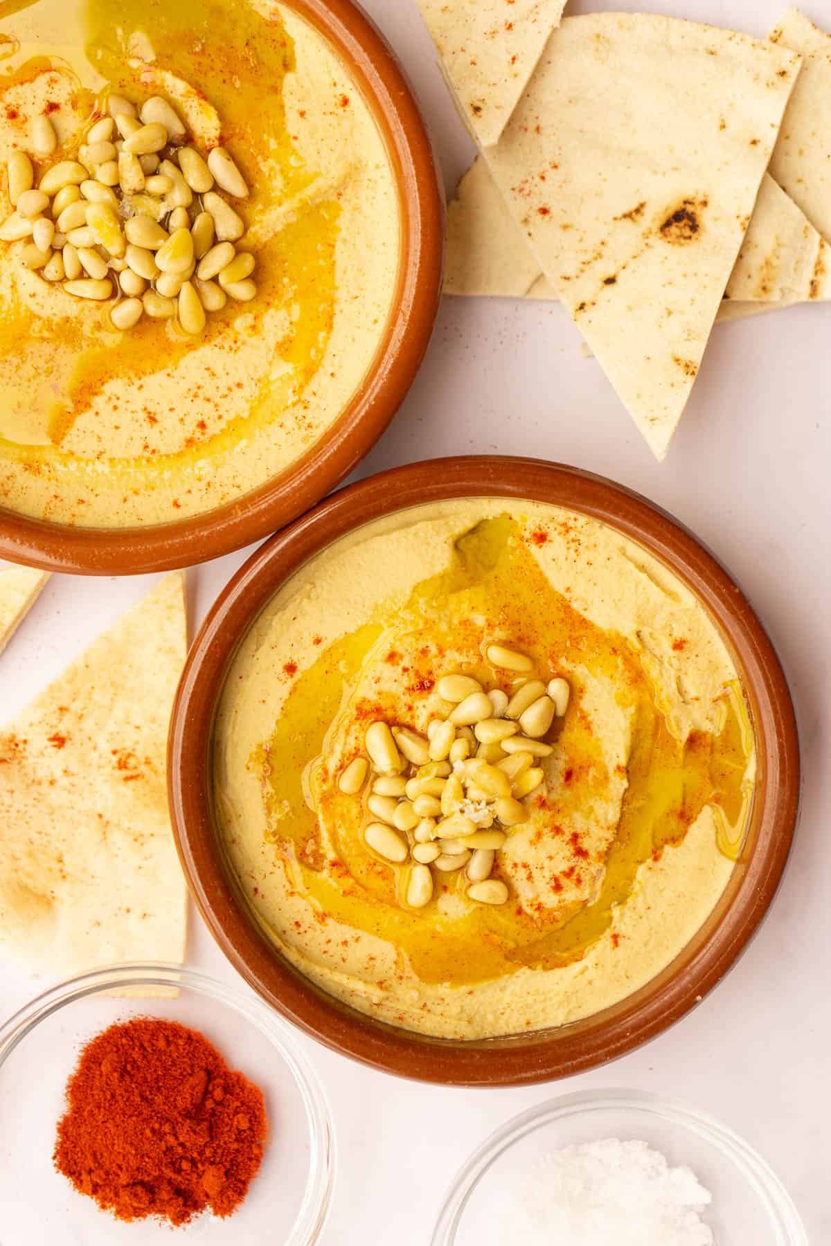 Two clay bowls of hummus topped with pine nuts, olive oil, and paprika, paired with pita bread.