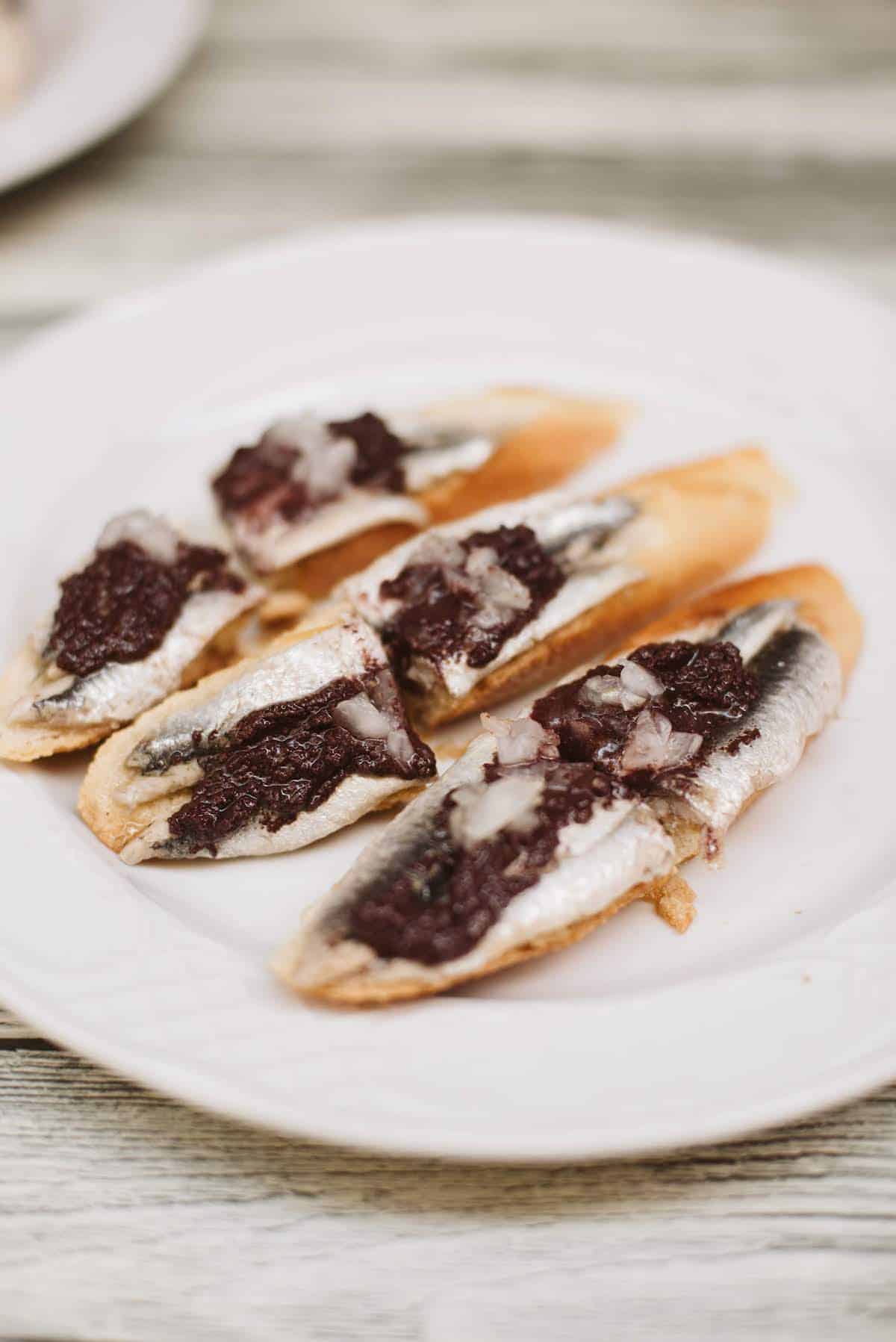 Plate of anchovies with black olive tapenade atop small slices of bread.