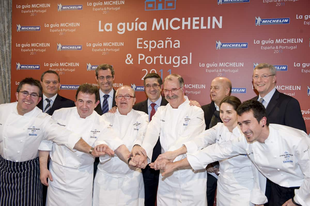 A group of people in chefs coats smiling and bumping fists, with a row of men in suits standing behind them.