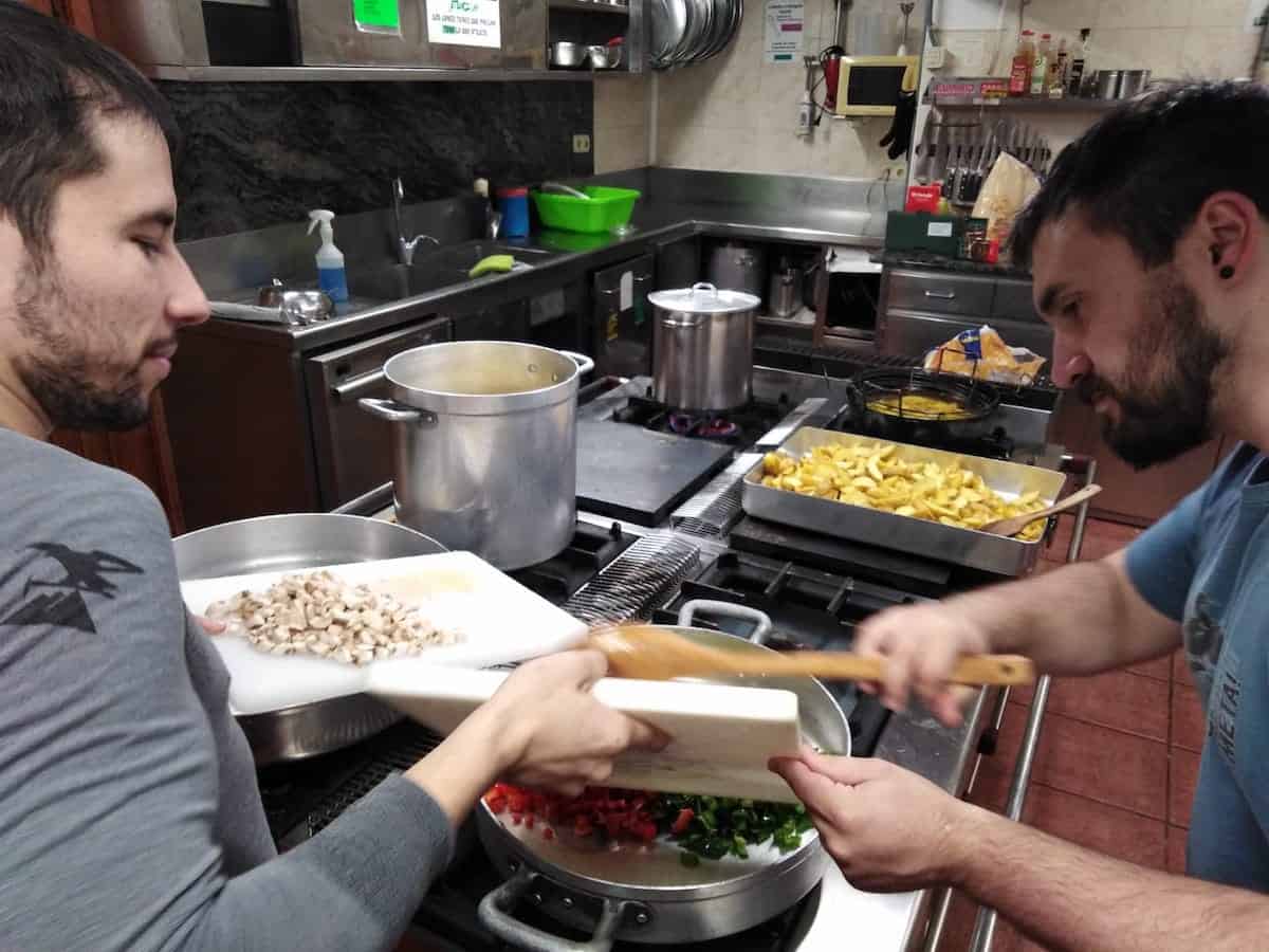 Two men adding chopped vegetables to a pot in a large industrial kitchen.