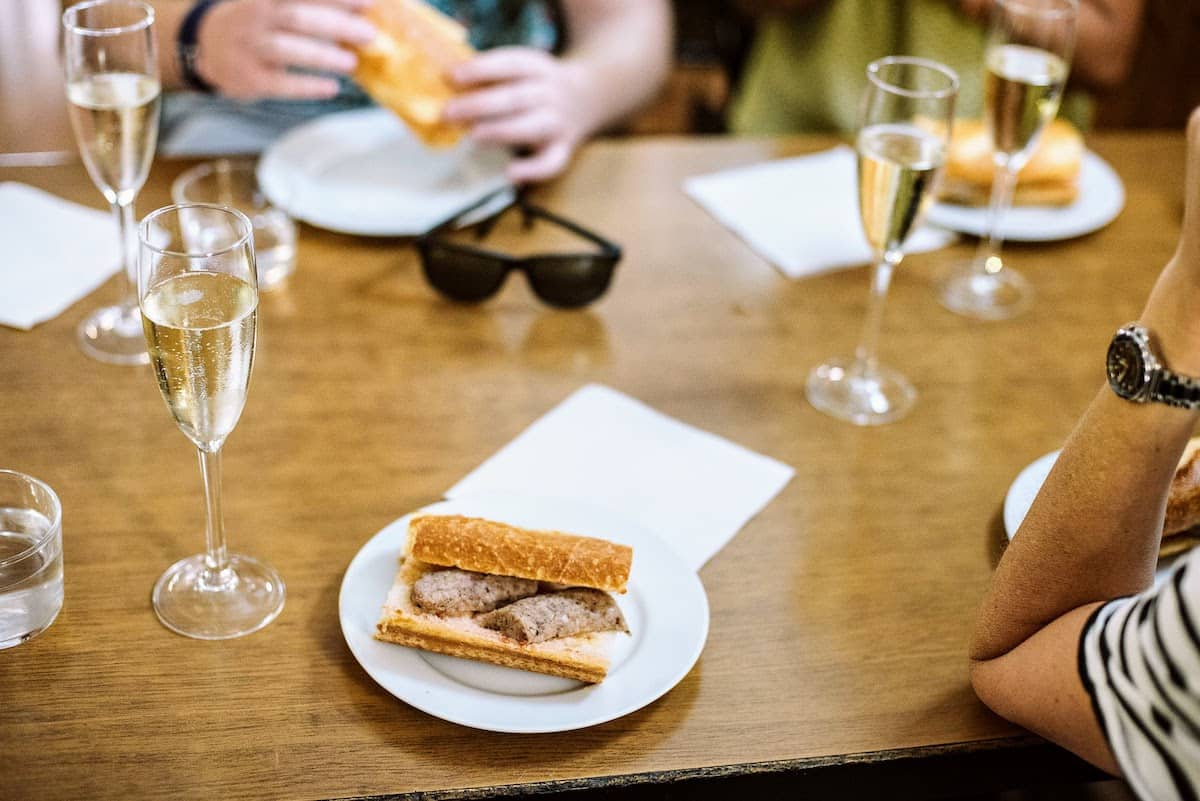 Slices of sausage in a bread roll on a white plate next to a glass of sparkling wine.