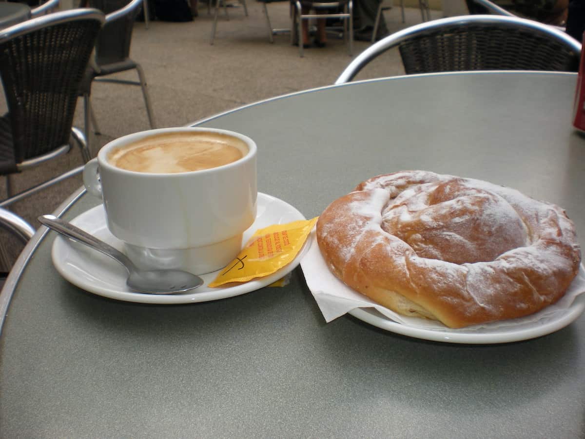 Cup of coffee with milk next to a spiral-shaped pastry dusted with sugar on an outdoor cafe table.