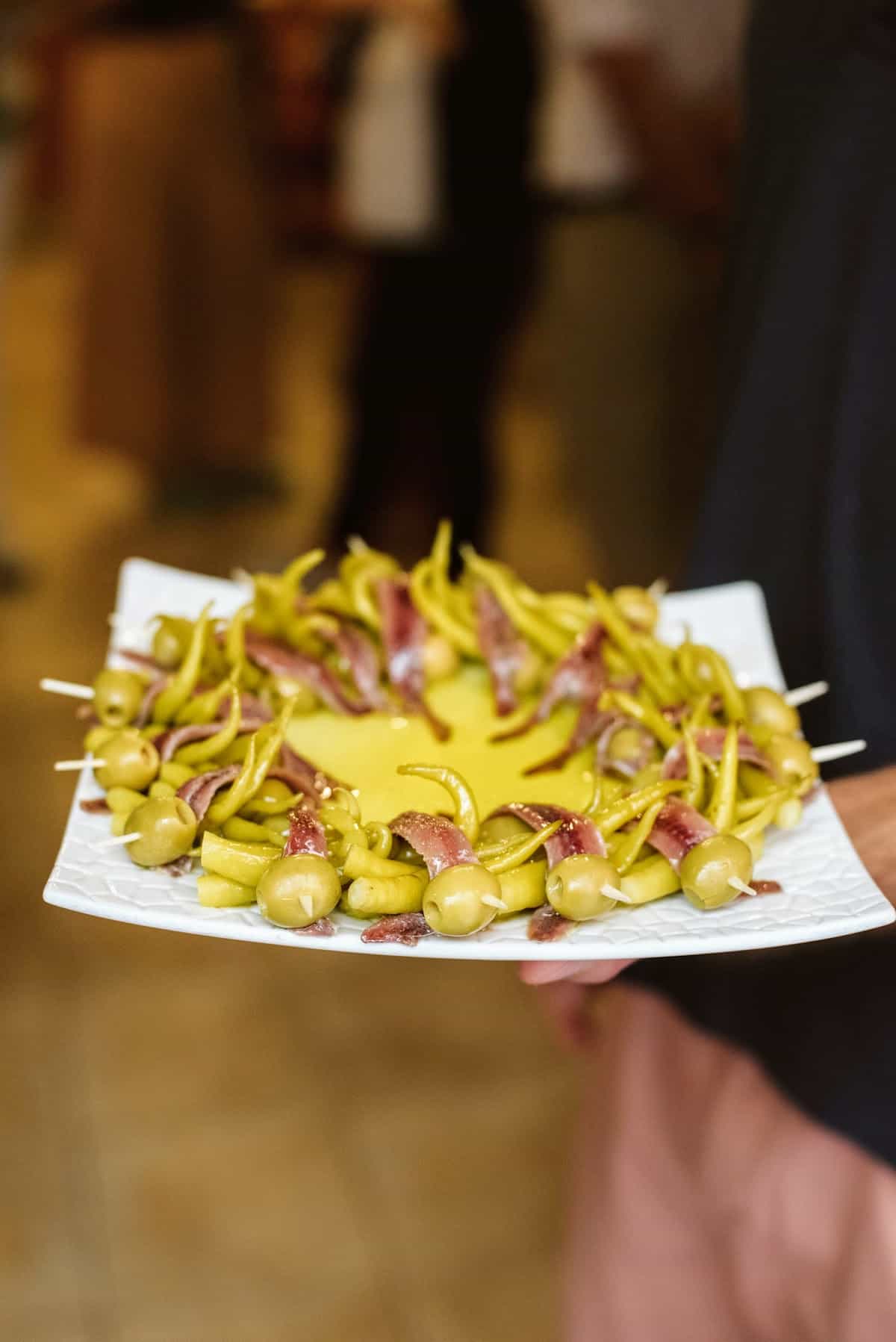 Skewers, or pintxos, of anchovies, peppers, and green olives arranged on a square plate.