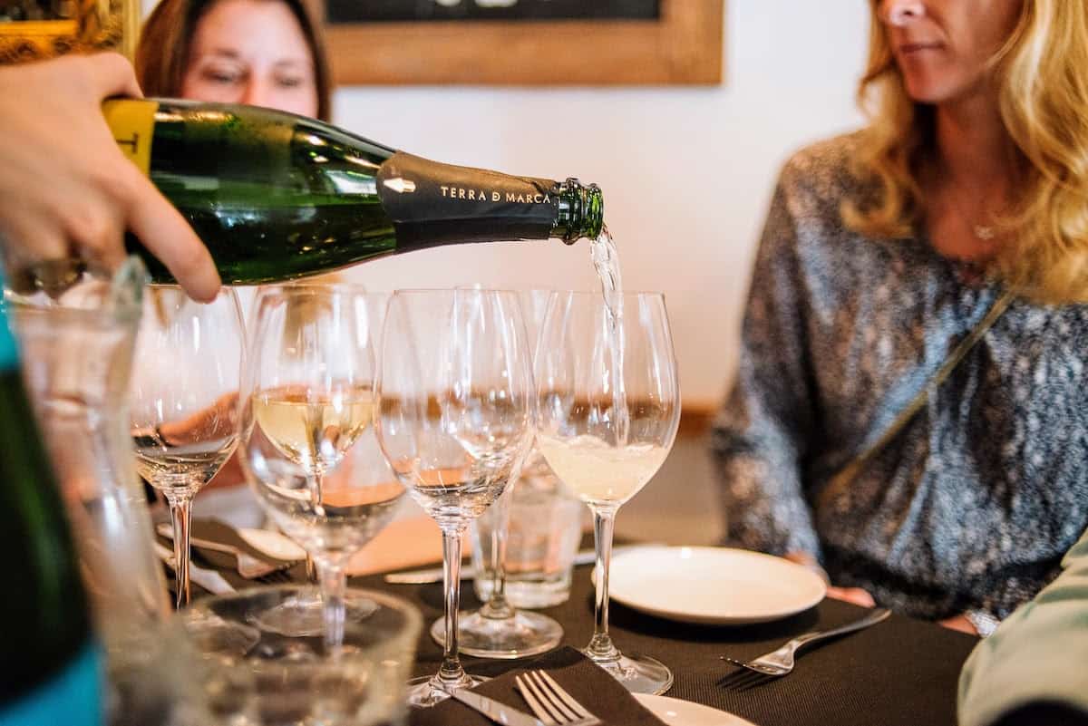 Person's hand pouring glasses of sparkling wine from a green bottle.